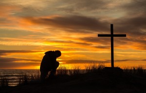 Beautiful sunset as man bows down to pray God before a cross.